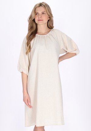 Femeie cu păr lung și blond, purtând o rochie bej, lejeră, lungime până la genunchi, cu mâneci bufante trei sferturi, stând în picioare pe un fundal alb.