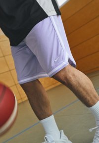 Lilla basketball shorts med hvide striber, lavet af åndbart stof. Har et Nike swoosh-logo. Parres med hvide sokker og sneakers.