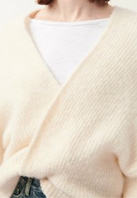 Cream-colored, soft knitted sweater with a V-neck design, featuring a textured pattern and an oversized fit, layered over a white shirt.