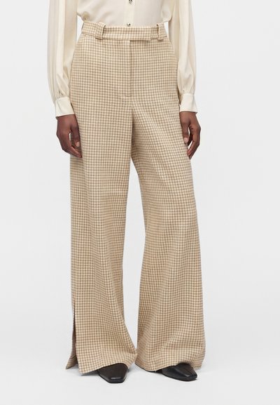 Pantalon beige à jambes larges avec un petit motif à carreaux, fentes devant aux ourlets, porté avec un chemisier crème et des chaussures noires à bout carré.