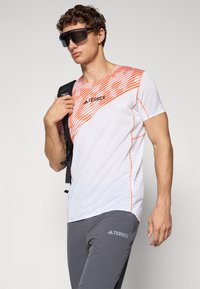 White short-sleeve shirt with orange diagonal stripes and logo; paired with gray pants featuring a logo; model carrying a backpack.