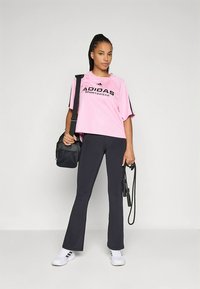 Pink cropped t-shirt with black "ADIDAS SPORTWEAR" logo, paired with dark leggings and white sneakers. Carrying a black bag and a jump rope.