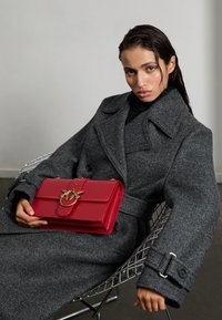 Červená kožená kabelka s kovovým zlatým zapínaním, so štruktúrovaným dizajnom a predným uzatváraním, držaná proti textúrovanej šedej bunde na drôtenej stoličke.