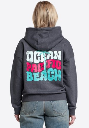 Osoba o krótkich blond włosach ubrana w ciemną bluzę z kapturem z kolorowym napisem "Ocean Pacific Beach" i grafiką palmy na plecach, zestawioną z dżinsami.