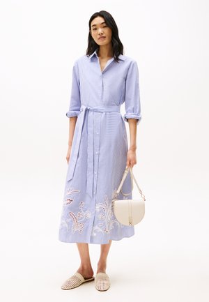 Abito a camicia a righe blu e bianche con vita regolabile, ricami floreali all'orlo, chiusura a bottoni e maniche a tre quarti. Borsa e scarpe beige.