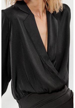 Kobieta ubrana w czarną, długorękawową bluzkę kopertową z głębokim dekoltem w kształcie litery V oraz lekko połyskującą, gładką fakturą tkaniny.