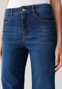 Primo piano di jeans in denim blu scuro a vita media con cuciture arancioni, indossati con una canotta bianca a coste, che mostrano le tasche anteriori e il dettaglio del bottone.