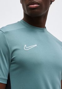 Άνδρας που φοράει πετρόλ αθλητική μπλούζα με λευκό λογότυπο Nike swoosh στο αριστερό μέρος του στήθους, εμφανιζόμενος από τον λαιμό μέχρι τη μέση του κορμού.