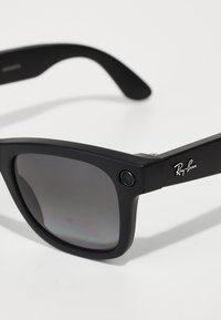 Schwarze Ray-Ban Sonnenbrille mit mattem Kunststoffrahmen, dunklen Verlaufsgläsern, metallenen Markenlogo am Bügel und einer kleinen Kamera, die nahe dem Scharnier integriert ist.