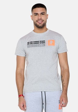 Homme portant un t-shirt gris clair avec le texte "WE ARE COMBAT SPORTS" et un logo de lion orange sur la poitrine, souriant à la caméra.