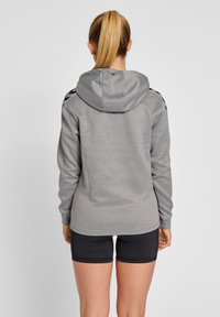 Sweat à capuche gris avec capuche, présentant des accents noirs sur les épaules. Le matériau semble texturé, porté avec un short noir et une finition lisse.