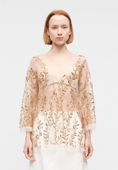 Διαφανές μπεζ μπλουζάκι με V-λαιμόκοψη, διακοσμημένο με κεντήματα φύλλων χρυσού και μακριά μανίκια, φορεμένο πάνω από λευκή φούστα με παρόμοια κεντήματα.