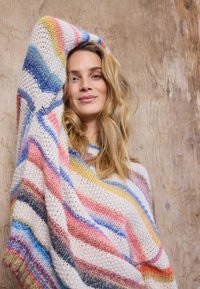 Mladá žena s vlnitými blond vlasy má na sobě pestrobarevný pruhovaný pletený svetr, zvedá jednu ruku a stojí před dřevěným pozadím.