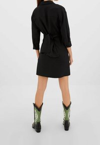 Vestido negro abotonado con cintura ajustada, mangas largas y corte recto. Combinado con botas vaqueras verdes y negras con detalles decorativos.