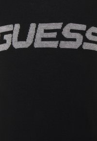 Schwarzer Strickpullover mit einem großen, auffälligen, weißen "GUESS"-Logo auf der Vorderseite. Der Stoff hat eine glatte Textur und ein körpernahes Design.