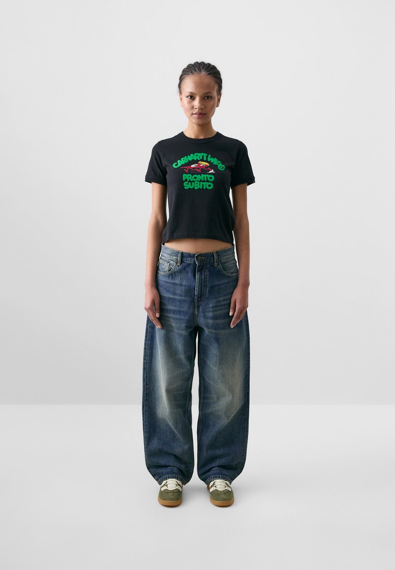 Black cropped t-shirt with green graphic text, paired with light-wash, oversized denim jeans and green-and-gold sneakers.