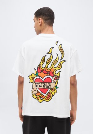 Άνδρας με την πλάτη, φοράει λευκό μπλουζάκι με πολύχρωμη απεικόνιση φλεγόμενης καρδιάς και λουλουδιών, με το κείμενο "PUMA" στην πλάτη.