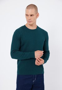 Teal gebreide sweater met een gestructureerd kabelpatroon, ronde hals en ribbelmanchetten. G gedragen met spijkerbroeken.