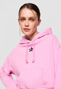 Felpa rosa con tasca a marsupio, caratterizzata da testo bianco "Tommy girl" e una stella nera in contrasto. Realizzata in tessuto morbido con cordini.