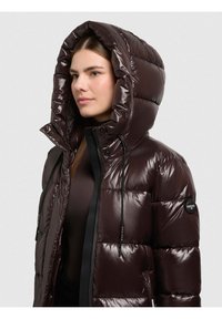 Sieviete valkā tumši brūnu spīdīgu polsterētu jaku ar lielu kapuci un melniem aukliņām, nedaudz paskatījusies pa kreisi.