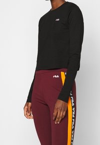 Svart långärmad crop top med en liten vit logotyp. Matchas med bourgognelila tights med orange och vita sidodekorationer.