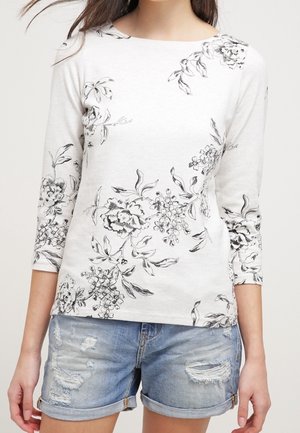 Kobieta w białym topie z czarnym kwiatowym nadrukiem i rękawem do łokcia oraz w jasnoniebieskich dżinsowych szortach z efektami przetarć.