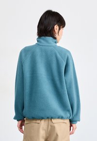 Pull en polaire turquoise avec un col montant, des manches raglan et une finition douce et texturée. Équipé d'un ourlet et de poignets élastiques.