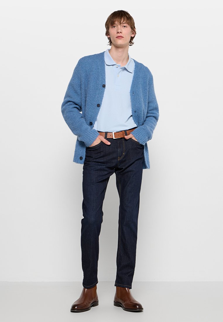 Hellblauer, kuscheliger Cardigan über einem blassblauen Poloshirt, dunklen Jeans und braunen Leder-Stiefeletten. Mit Knopfdetails und lockerer Passform.