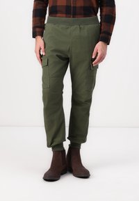 Zelené cargo kalhoty s elastickým pasem, zúženými manžetami a bočními kapsami, nošené s hnědými kotníkovými botami. Bavlněná směs, hladká textura.