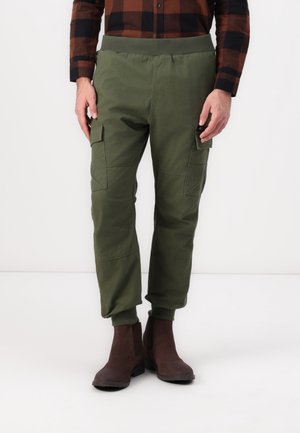 Gröna cargo-byxor med elastisk midja, avsmalnande ärmslut och sidofickor, bärs med bruna ankelboots. Blandning av bomull, slät textur.