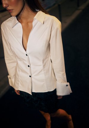 Valkoinen napapaita, jossa on syvä V-kaula-aukko, pitkät hihat ja mustat napit. Yhdistetty teksturoidun mustan hameen kanssa. Tumma tausta.