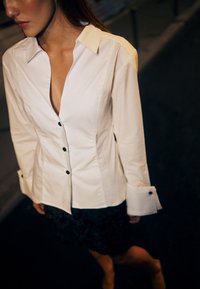 Balti mygtukų marškiniai su giliu V formos iškirpimu, ilgomis rankovėmis ir juodais mygtukais. Sukomplektuoti su tekstūruota juoda sijonu. Tamsus fonas.