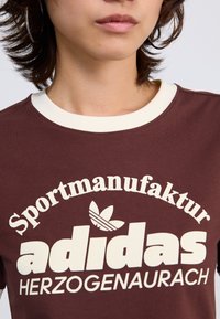 Brown T-shirt with white ribbed neckline, featuring "Sportmanufaktur" and "adidas" text in bold typography and logo graphic at center.