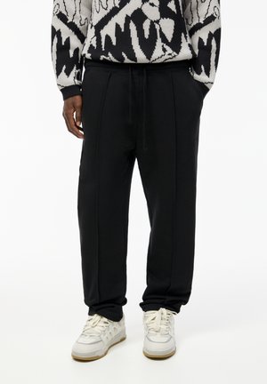 Persona vestida con pantalones de chándal negros con cordón, suéter con patrón en blanco y negro, y zapatillas blancas, de pie frente a un fondo blanco.
