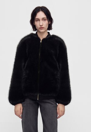 Young person with short dark hair wearing a black faux fur zip-up jacket and dark gray pants, standing against a plain light background.
