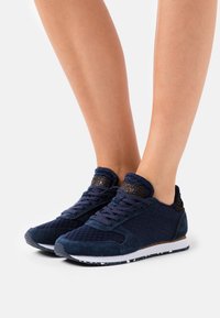 Baskets bleu marine avec une tige en mesh texturée et des accents en daim. Présentent un col en tissu foncé à motifs et une semelle blanche tachetée.