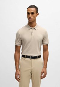 Beige polo skjorta med krage och korta ärmar, tillverkad av texturerad tyg. Har en subtil logo på bröstet och är kombinerad med ljusbeige byxor.