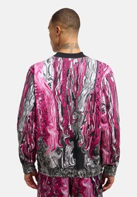 Fargerik jakke med glidelås som har svirrende mønstre i rosa, svart og hvit, med ribbestrikket krage og elastisk midje.