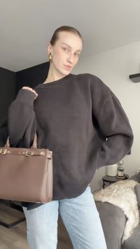 Bolso de cuero marrón con un diseño estructurado, herrajes en tono dorado, textura suave; llevado con un sudadera oversized negra y jeans de un azul claro.