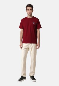 Bordeauxfarbener Baumwoll-T-Shirt mit kurzen Ärmeln, Rundhalsausschnitt und Logo auf der Vorderseite. Kombiniert mit hellbeigen Hosen und schwarzen Sneakern.