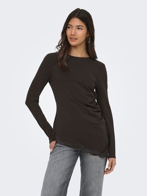Ung kvinne iført en svart langermet topp med blonder i kanten på kanten, kombinert med lys grå jeans, står og ser til siden.
