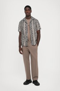 Kortermet skjorte med slangeprints i lysegråtoner, sammen med beige bukser. Sorte joggesko, enkel t-skjorte med rund hals under.