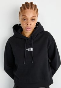 Žena s vrkôčkami a kruhovými náušnicami, oblečená v čiernej mikine značky The North Face, na plain svetlom pozadí.