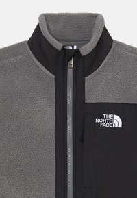 Giacca in pile grigia con un pannello in nylon nero, colletto alto, zip intera e tasca sul petto a sinistra. Presenta un logo bianco sulla tasca.