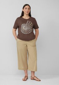 Bruine t-shirt met korte mouwen met een grafische afbeelding van een witte en gele nautilusschelp, gecombineerd met lichtbeige wijde broek en bruine sandalen.