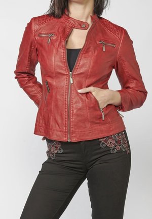 Donna che indossa una giacca di pelle rossa con tasche con cerniera, top nero e pantaloni neri con ricamo floreale rosso e bianco sui fianchi.
