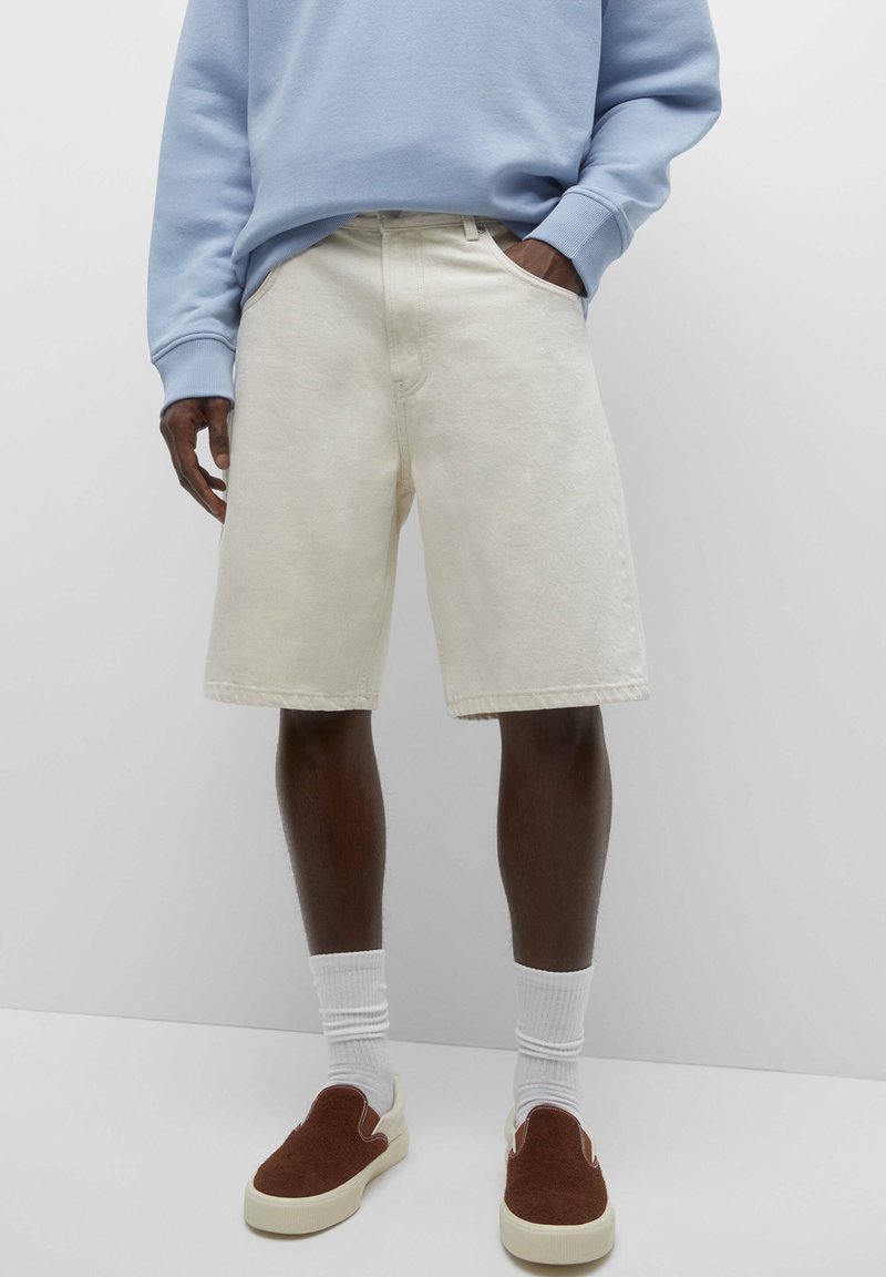 Shorts en denim beige avec poches avant et coupe droite, associés à des chaussures marron à enfiler et des chaussettes blanches, sur un fond gris.