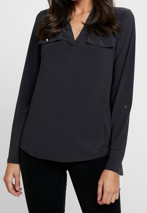 Žena v dlouhém černém topu s dvěma kapsami na hrudi a výstřihem do V, v kombinaci s černými kalhotami.