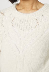 Leichter, cremefarbener Pullover mit strukturiertem Strickmuster und Lochdetails, die ein Chevron-Design am Ausschnitt bilden.