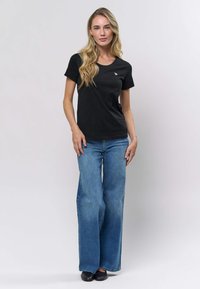 Črna kratka majica s kratkimi rokavi in okroglim izrezom ter majhno logotipsko podrobnostjo, kombinirana z lahkimi modrimi širokimi kavbojkami in ravnimi črnimi čevlji.